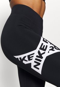 Czarne legginsy wykonane z elastycznego materiału, z dużą białą grafiką z napisem "NIKE" po boku, gładką teksturą i wysokim stanem.