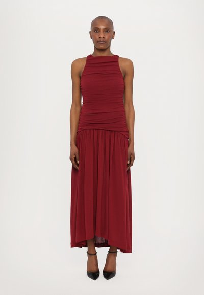 Bordeauxrotes ärmelloses Kleid mit gerafftem Oberteil und fließendem Rock. Glatter Stoff, knielanger Saum und schwarze High Heels.