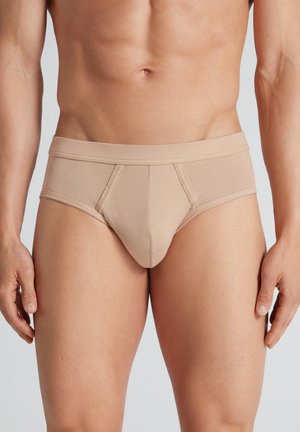 Männlicher Torso trägt beige Slips, steht mit entspannten Armen seitlich am Körper vor einfachem Hintergrund.