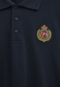 Polo blu navy con tre bottoni e uno stemma ricamato che presenta una corona, una corona di alloro e bandiere colorate con la scritta "Polo Club".