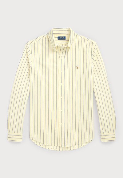 Knappskjorte i lys gul med blå vertikale striper. Har krage, lange ermer, avrundet nederkant og en liten logo nær brystet.