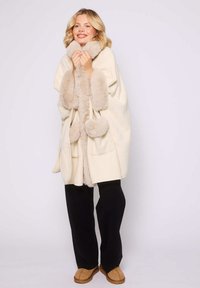 Poncho crema con acentos de pelo de imitación beige, que presenta un cuello ancho, lazos frontales y detalles de pompones redondos, usado sobre pantalones negros.