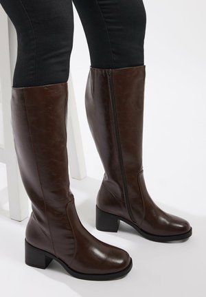 Brązowe skórzane kozaki do kolan z kwadratowym noskiem, gładką teksturą, zamkiem błyskawicznym z boku oraz słupkowym obcasem. Wyeksponowane obok czarnych legginsów.