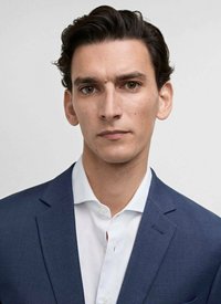 Man med mörkt, vågigt hår, iförd en marinblå blazer med ett subtilt mönster och en vit skjorta. Han har ett neutralt uttryck och en jämn hy.