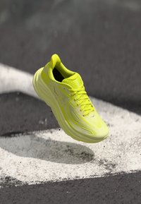 Neon-gelbe Laufschuhe mit einer strukturierten Oberseite, reflektierenden Akzenten, gepolsteter Sohle und leichtem Design, in der Luft über einer weißen Linie dargestellt.