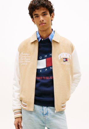 Junger Mann trägt eine beige-weiße Collegejacke über einem marineblauen Tommy Jeans Sweatshirt und hellblauen Jeans, steht vor einem weißen Hintergrund.