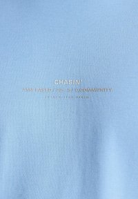 Světle modrý bavlněný sweatshirt s embosovaným textem: "CHASIN' AMS BASED / 22-5 / C.COMMUNITY AUTHENT TECH DENIM." Hladký povrch.