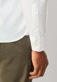 Camisa blanca de manga larga con puños abotonados, que presenta una textura suave y un ajuste entallado; los botones visibles son de color perla.