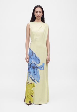 Frau, die ein ärmelloses, blassgelbes bodenlanges Kleid mit großem blau-grünem Blumenmuster trägt, steht vor einem schlichten weißen Hintergrund.