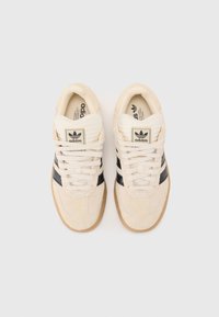 Beige suede sneakers with black accents, white laces, and a textured rubber sole. Features classic branding on the tongue and side.