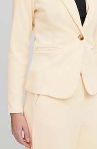 Svetlobéžový priliehavý blazer s ribbovanou textúrou, jedným gombíkom a bočnými vreckami. Dizajn obsahuje klopový golier a zúžený strih.