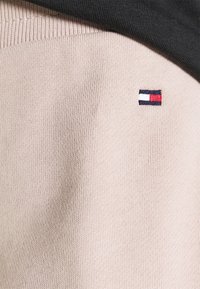 Hellbeige gestrickter Stoff mit einem kleinen gestickten Logo und roten, weißen sowie marineblauen Streifen. Oben mit dunkelgrauem Stoff überlagert.
