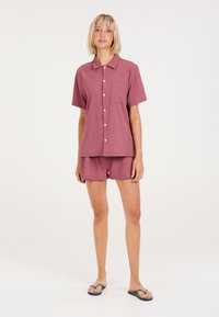 Top de manga corta con cuello y pantalones cortos a juego en tejido granate texturizado. Bolsillo frontal en la parte superior; modelo vistiendo chanclas.