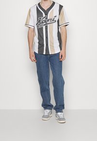 Camisa estilo béisbol en blanco, gris y beige con rayas verticales, mangas cortas y un logo negro. Combinada con jeans de mezclilla azul y zapatillas grises.