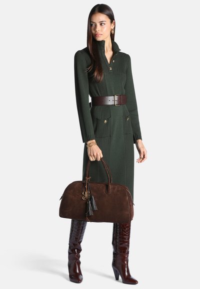 Femme portant une robe ceinturée vert foncé, tenant un sac à main en cuir marron et portant des bottes hautes brillantes marron à motif crocodile.