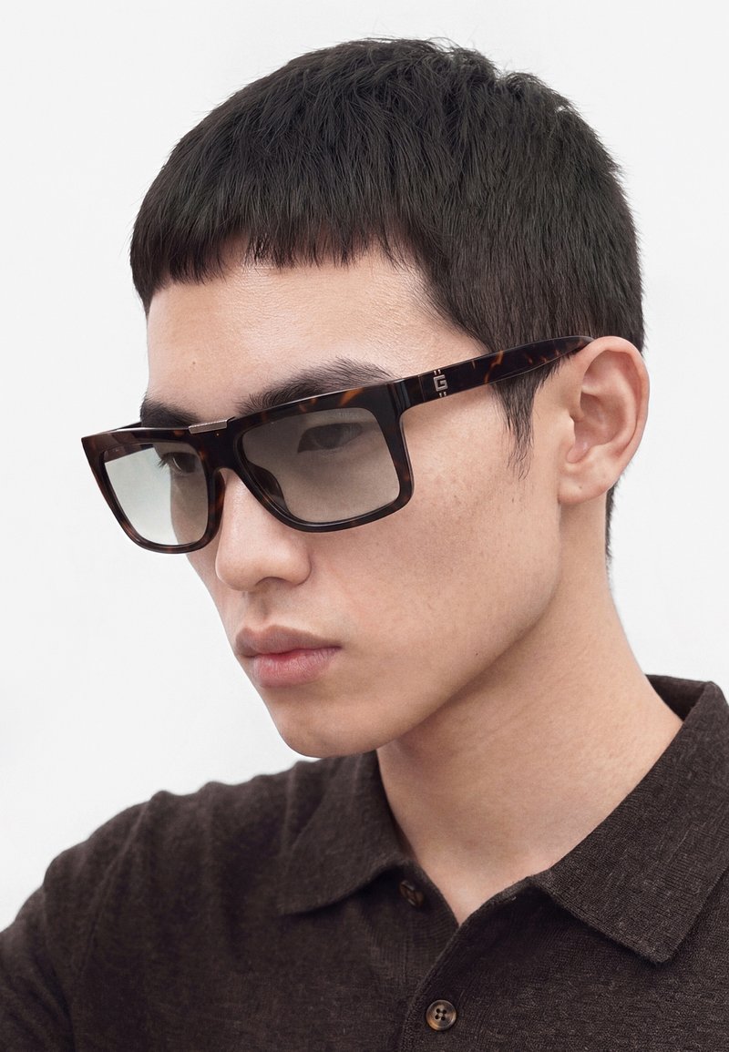 Jeune homme aux cheveux courts et noirs portant de grandes lunettes de soleil rectangulaires écaille de tortue et une chemise sombre boutonnée, regardant légèrement vers le bas.