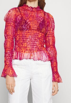 Donna che indossa una camicetta trasparente a maniche lunghe arricciate con motivi floreali rosa e arancioni, sopra un reggiseno viola, abbinata a pantaloni bianchi a vita alta.