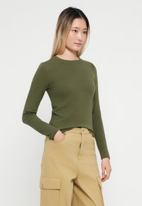 Tricou cu mâneci lungi, ajustat, de culoare verde măsliniu, confecționat dintr-un material moale; decolteu rotund și textură netedă, asociat cu pantaloni cargo bej.