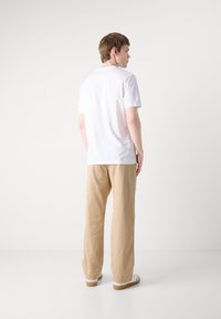 Tricou alb cu mânecă scurtă, cu croială dreaptă, asortat cu pantaloni bej cu croială lejeră și adidași albi cu talpă maro, prezentat din spate.