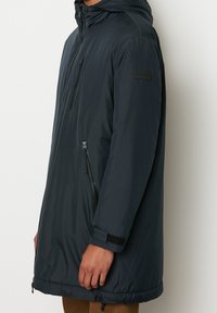 Parka lunga blu navy con cappuccio, apertura frontale con zip, due tasche laterali con zip e polsini con velcro. Texture liscia, materiale leggero e dettagli minimi.