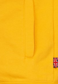 Žltá mikina so klokaním vreckom, rebrovaným lemom a malým britským vlajkovým akcentom na boku. Mäkká textúra, neformálny dizajn.