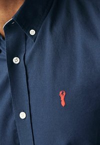 Chemise bleu marine à boutons, fabriquée en tissu texturé, avec un logo de cerf brodé en rouge sur la poitrine gauche et des boutons blancs.