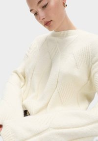 Suéter de punto color crema con un patrón geométrico texturizado, cuello redondo acanalado y mangas anchas. Material suave y acogedor.