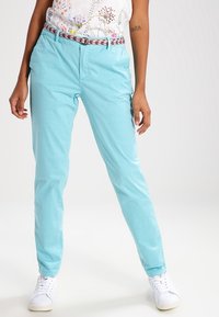 Kobieta ubrana w jasnoniebieskie spodnie z różowym wzorzystym paskiem, białą kwiecistą bluzkę i białe sneakersy, stojąca na tle jednolitego jasnego tła.