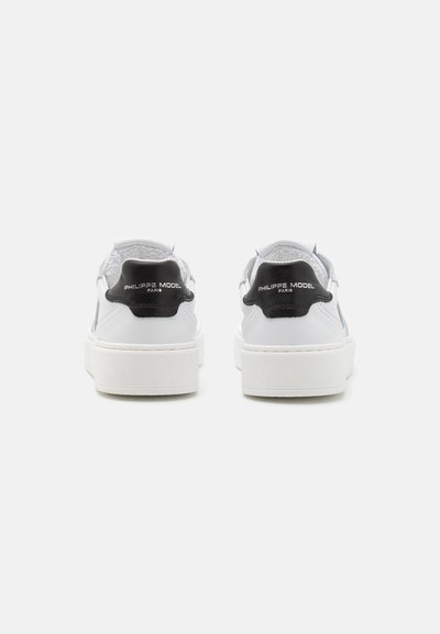Baskets en cuir blanc avec un dos texturé en noir, dotées d'une semelle plate et d'un détail logo argenté sur le talon. Design simple et moderne.