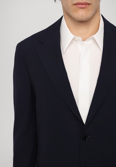 Giacca di suit in tessuto navy con revers a cresta e un singolo bottone nero, indossata su una camicia bianca con colletto rigido.