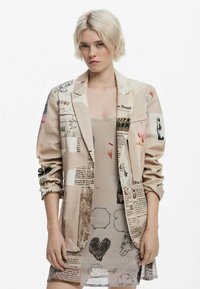 Beige blazer with mixed media prints including text, illustrations, and abstract designs. Smooth texture, single-breasted style with rolled sleeves.