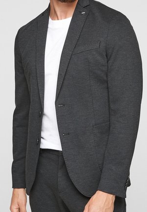Homme portant une veste de costume gris foncé cintrée et un pantalon assorti avec un t-shirt blanc uni en dessous.