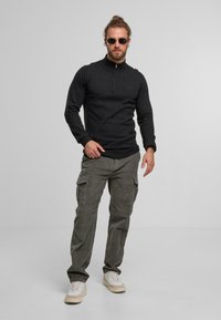 Sudadera negra con cremallera y cuello alto, combinada con pantalones cargo verdes oliva que tienen bolsillos laterales. El calzado son zapatillas beige.