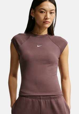 Kvinde iført en tætsiddende, kortærmet, ribstrikket lilla Nike-top og matchende bukser, står med hænderne i lommerne mod en ensfarvet baggrund.