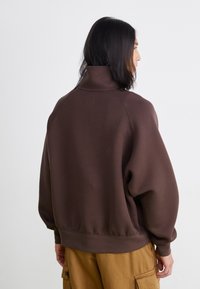 Brun sweatshirt med hög krage, löst passform och ribbade manschetter. Tyget verkar mjukt och texturerat, med en enfärgad design och inga synliga mönster.