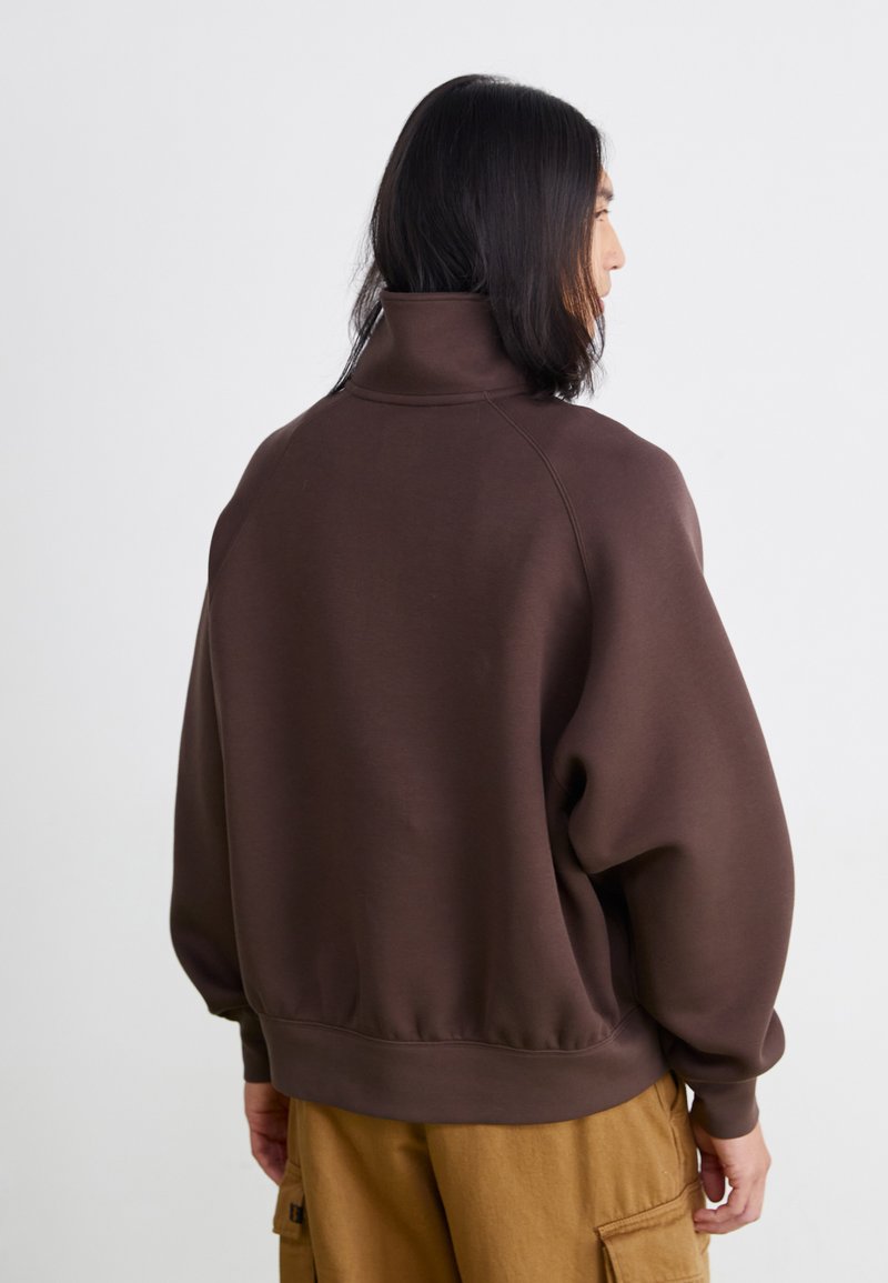 Sudadera marrón con un cuello alto, corte holgado y puños acanalados. La tela parece suave y texturizada, con un color sólido y sin patrones visibles.