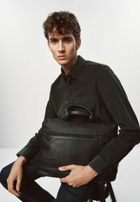 Czarna skórzana torba na laptopa o teksturowanej powierzchni, z zamkiem błyskawicznym i krótkimi uchwytami, trzymana przez modela w ciemnej koszuli.