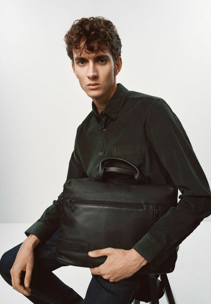 Czarna skórzana torba na laptopa o teksturowanej powierzchni, z zamkiem błyskawicznym i krótkimi uchwytami, trzymana przez modela w ciemnej koszuli.