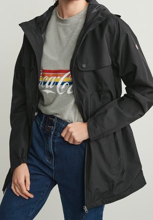 Personne portant une veste à capuche noire par-dessus un t-shirt gris avec le logo Coca-Cola et un jean bleu taille haute, ajustant la veste d'une main.