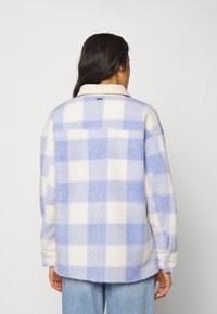 Vrouw met lang, donker haar, gekleed in een blauw-witte geruite fleece jas en lichtblauwe spijkerbroek, gezien vanaf de achterkant tegen een effen achtergrond.