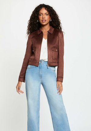 Mujer con chaqueta marrón con cremallera, camiseta blanca, vaqueros de pierna ancha de talle alto de color azul claro, con pelo rizado sobre un fondo liso.