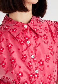 Blusa floral roja con botones de perlas brillantes y un cuello puntiagudo, llevada por una persona con cabello corto y oscuro.