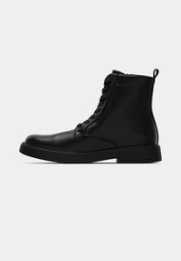 Bottine noire en cuir avec bout arrondi, fermeture à lacets à l'avant, languette à l'arrière et semelle plate sur fond blanc.