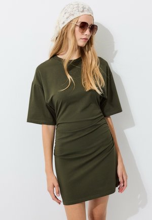 Femme aux longs cheveux blonds portant des lunettes de soleil, un foulard en dentelle blanche et une robe froncée à manches courtes vert olive, debout contre un mur blanc.