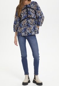 Blouse florale dans des tons marine, bleu et pêche ; présente des manches longues, une taille froncée et un tissu léger. Associée à un jean bleu et des bottes crème.