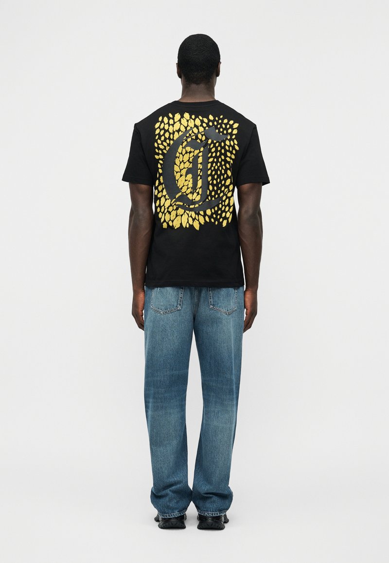 Άνδρας που φοράει μαύρο T-shirt με μεγάλο κίτρινο σχέδιο φύλλου και μαύρο στυλιζαρισμένο γράμμα στην πλάτη, σε συνδυασμό με μπλε τζιν και μαύρα παπούτσια.