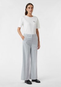 Witte t-shirt met korte mouwen en een ronde hals, gecombineerd met lichtgrijze wijde broek en zwarte instappers, staand op een effen achtergrond.