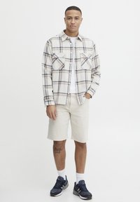 Gekruiste shirt in crème, blauw en bruin; korte beige shorts; zwart-blauwe sneakers; effen wit T-shirt eronder; twee voorzakken.