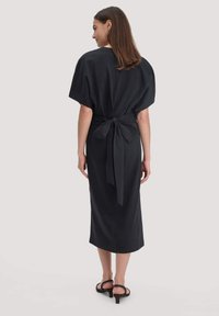 Frau, die mit dem Rücken steht, trägt ein schwarzes Midi-Kleid mit weiten Ärmeln und einer gebundenen Schleife am Rücken, kombiniert mit schwarzen High Heels.