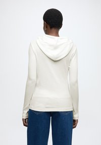 Feh�ér hosszú ujjú kapucnis felső kenguru zsebbel, bordázott textúrával és állítható húzózsinóros kapucnival. Kék, bő szárú farmerrel párosítva.